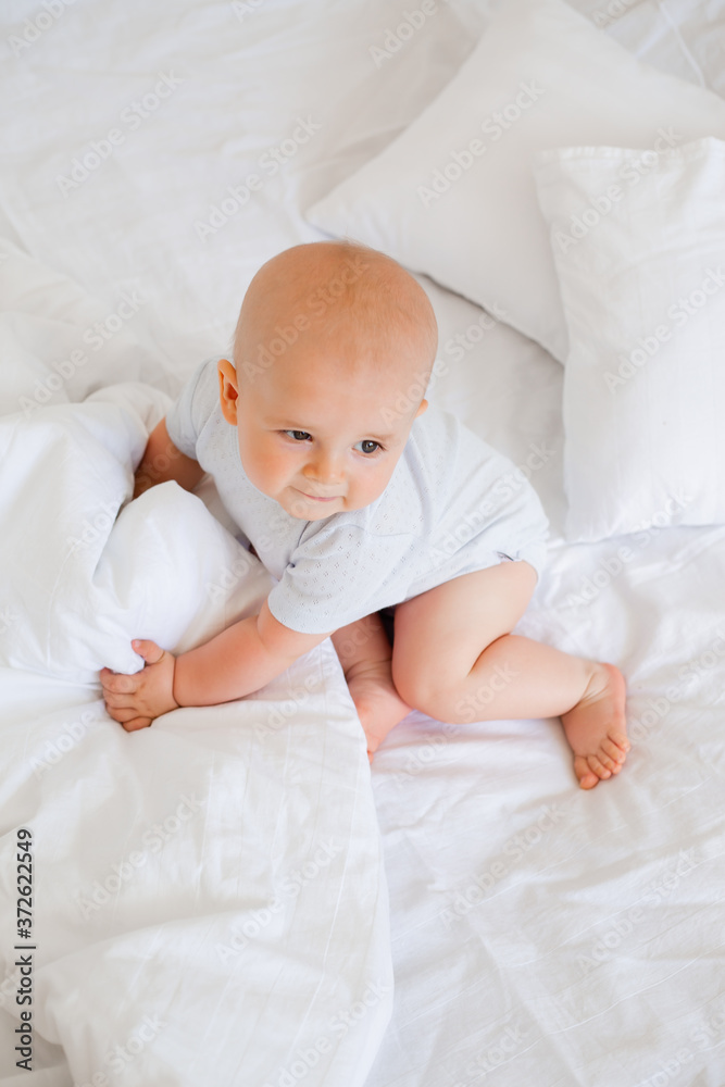 Cute baby boy 6 months smiling in a white bodysuit sitting on a bed on white bedding Cute baby boy 6 months smiling in a white bodysuit sitting on a bed on white bedding