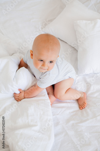 Cute baby boy 6 months smiling in a white bodysuit sitting on a bed on white bedding