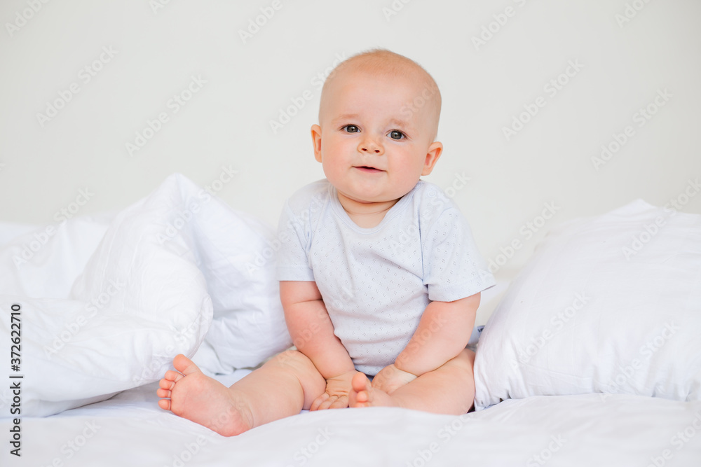 Cute baby boy 6 months smiling in a white bodysuit sitting on a bed on white bedding Cute baby boy 6 months smiling in a white bodysuit sitting on a bed on white bedding