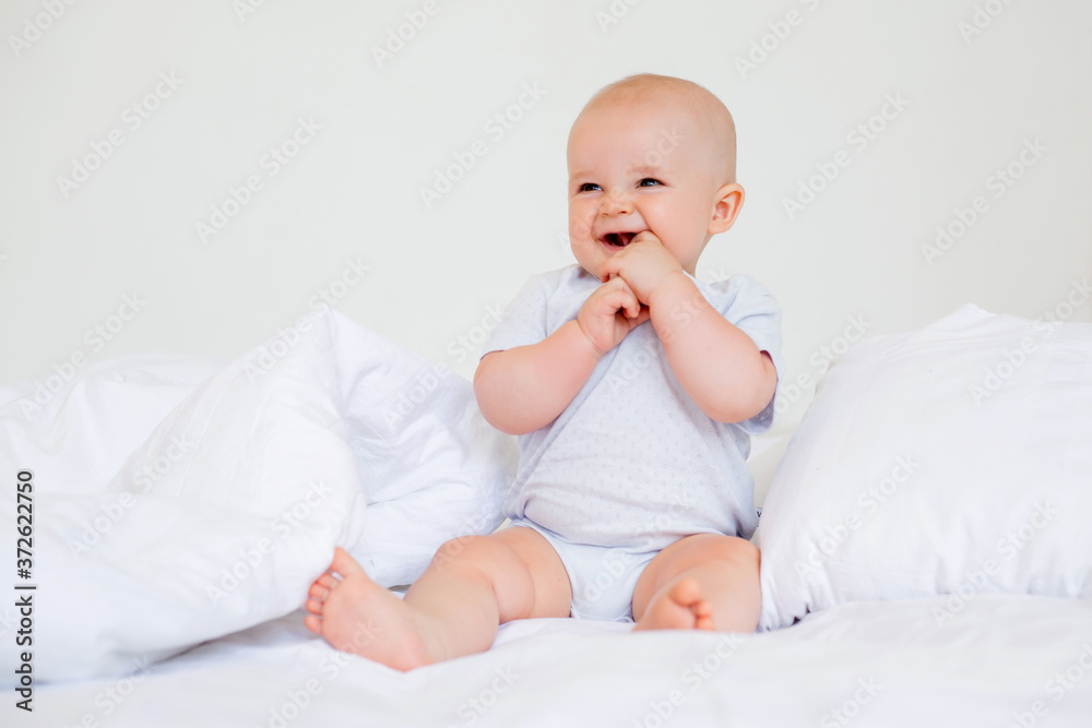 Cute baby boy 6 months smiling in a white bodysuit sitting on a bed on white bedding Cute baby boy 6 months smiling in a white bodysuit sitting on a bed on white bedding