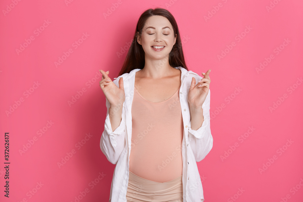 Young beautiful pregnant girl expecting baby isolated over pink background gesturing finger crossed and smiling with hope, keeps eyes closed, dresses casual outfit. Young beautiful pregnant girl expecting baby isolated over pink background gesturing finger crossed and smiling with hope, keeps eyes closed, dresses casual outfit.