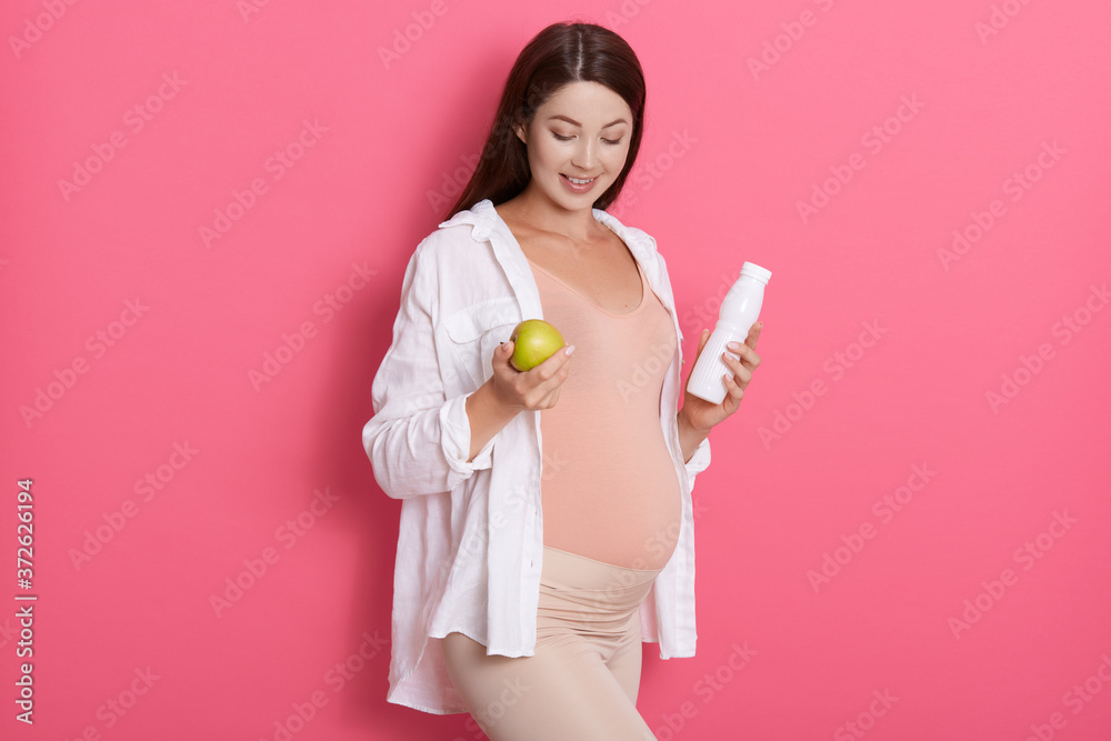 Pregnant woman with pretty stomach holding green apple and bottle of milk, expectant mother looking down, posing isolated over rosy background, wearing white shirt.