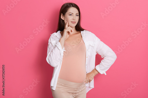 Pensive pregnant woman wearing white shirt and leggins, dark haired female looking aside with finger on her cheek, standing isolated over rosy background.