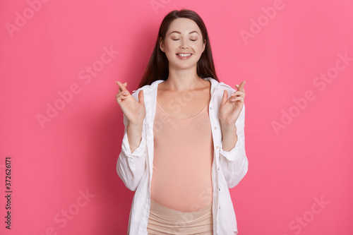 Young beautiful pregnant girl expecting baby isolated over pink background gesturing finger crossed and smiling with hope  keeps eyes closed  dresses casual outfit.