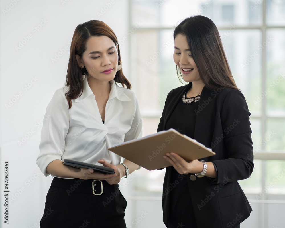 Two female office workers in business suit discuss about their work with tablet and file in hand in business building. Businesswoman talking and poitning on tablet. Business stock photo Two female office workers in business suit discuss about their work with tablet and file in hand in business building. Businesswoman talking and poitning on tablet. Business stock photo