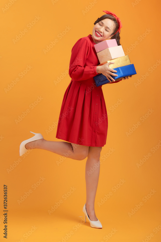 Smiling young Asian lady holding gift boxes isolated over orange background. Smiling young Asian lady holding gift boxes isolated over orange background.