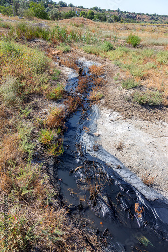 Spilled streams of liquid crude oil flow down drainage ditch into public body of water. Environmental disaster Oil pollution of environment from obsolete technologies and ancient production equipment