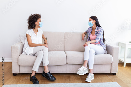 Two female friends in social distancing sitting on sofa in coronavirus pandemic time. Best friends having coffee together while separated by social distancing on sofa at home