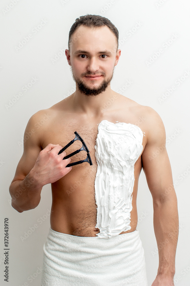 Fototapeta premium Young caucasian man with beard holds razor shaves his chest with white shaving foam on white background. Man shaving his torso