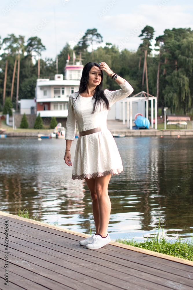A woman walks along the embankment along the river. A brunette in a white dress.