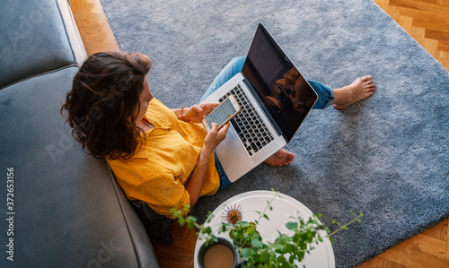 Obraz na płótnie young girl in a yellow shirt woman working on a laptop at home using a smartphone in the living room on the carpet, distant work and education w salonie