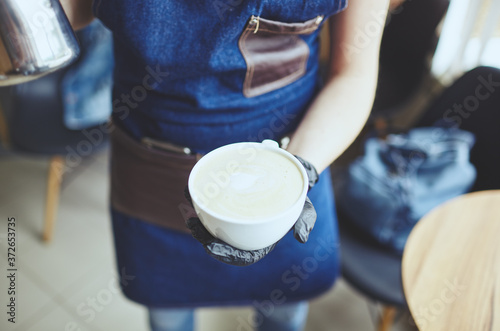 Female holds a milk jug and a cup of coffee, makes cappuccino, pouring whipped milk into espresso.Blurred image, selective focus