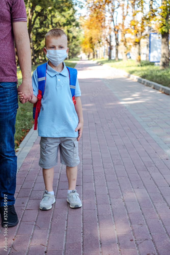 Portrait of a schoolboy in a protective mask in the park. Safety mask to coronavirus prevention.The father holding the first-grader's hand. Education. Portrait of a schoolboy in a protective mask in the park. Safety mask to coronavirus prevention.The father holding the first-grader's hand. Education.