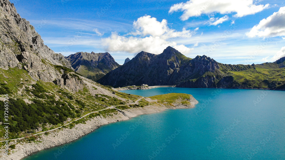 Aerial view of a path or trail without any people at Lünersee, Austria. You can see the turquoise lake, green grassland and the rocky tops of the mountains.