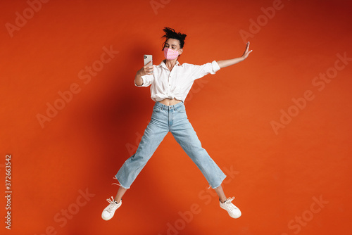 Image of young joyful woman jumping and taking selfie on cellphone
