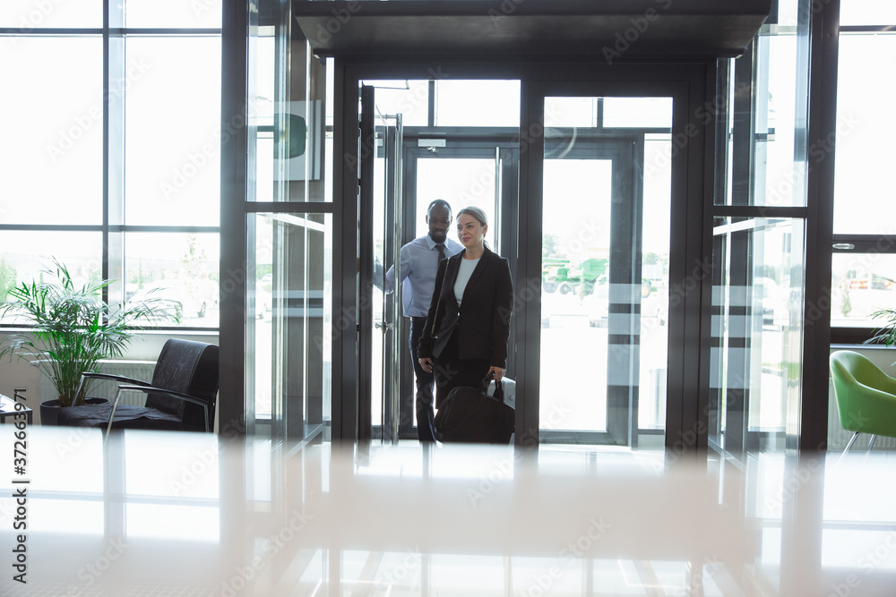 Greeting. Meeting of young business partners after arriving to end point of business trip. Man and woman walking against glass wall background of modern building. Concept of business, finance, ad. Greeting. Meeting of young business partners after arriving to end point of business trip. Man and woman walking against glass wall background of modern building. Concept of business, finance, ad.