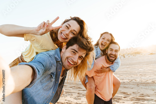 Image of young people smiling and taking selfie while walking on beach