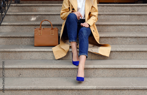 Street fashion outfit, model in blue jeans coat and brown handbag in high heel shoes sitting on the beige stair, outdoor