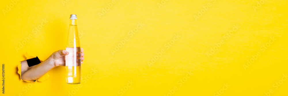 Female hand holding a glass bottle with water on a bright yellow background. Banner. Female hand holding a glass bottle with water on a bright yellow background. Banner.