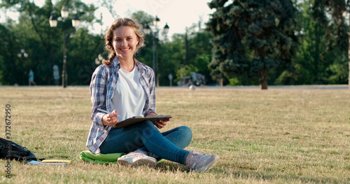 A student girl in a plaid shirt sits on the grass in the park and works on the tablet, prepares for the classes, looks at the camera and smiles.
