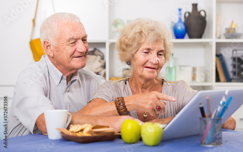 Happy elderly pair using laptop sitting at table at home.