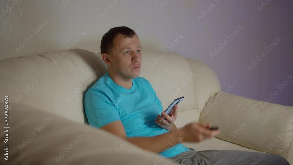 Man sits on the couch in the evening reading messages on the phone and watches the news on TV Man sits on the couch in the evening reading messages on the phone and watches the news on TV