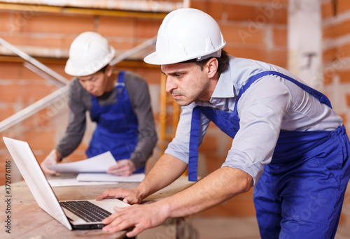 Portrait of two builders reading plan of brick building using laptop