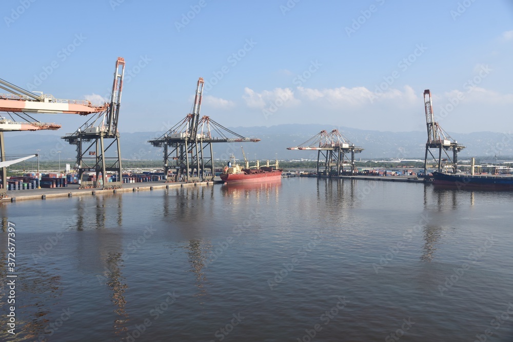 View on Kingston Harbour in Jamaica is the seventh-largest natural harbour in the world. In the background are gantry cranes and red general bulk cargo vessel and blue container ship.