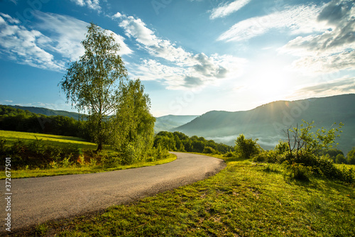 asphalt road near glades and trees on a background of mountains. Sunbeams.