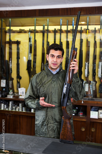 Portrait of man in hunting raincoat in gun store carefully picking out rifle