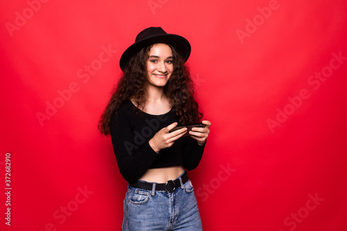 Cheerful joyful young woman playing games on smartphone and having fun over red background