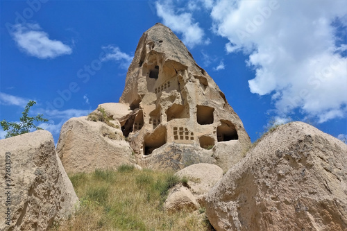 Ancient conical rock of Cappadocia against blue sky. Caves have been carved into the stone. In the foreground there are boulders, yellowed grass. Summer sunny day. Unesco heritage. Turkey