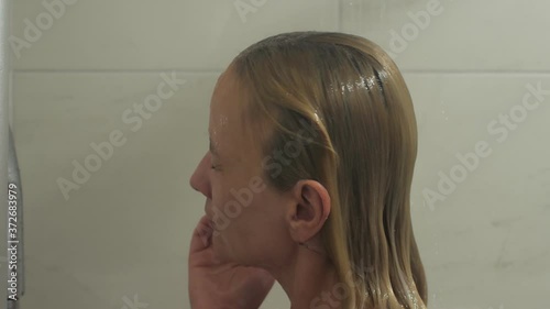 Naked young blonde takes a shower in a glass shower. She washes her hair. Slow motion shot.
