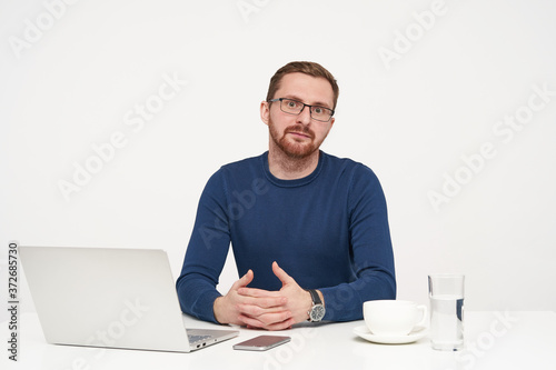 Puzzled young unshaved fair-haired man dressed in blue sweater looking surprisedly at camera and keeping his hands on tabletop, sitting over white background