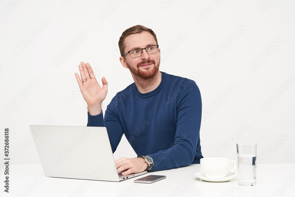 Handsome young unshaved fair-haired man dressed in blue sweater raising palm in hello gesture while looking aside, working with his laptop over white background Handsome young unshaved fair-haired man dressed in blue sweater raising palm in hello gesture while looking aside, working with his laptop over white background