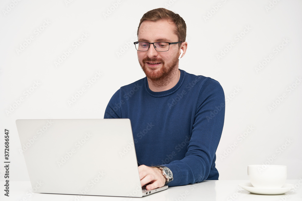 Positive young pretty bearded male with earphones keeping hands on keyboard and smiling gladly while typing text, isolated against white background