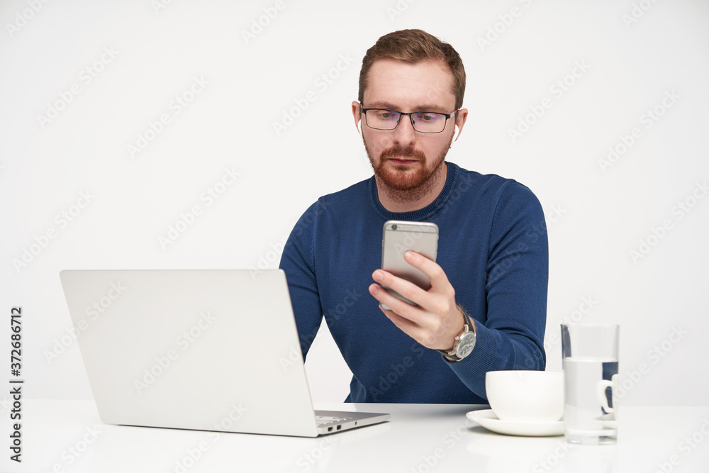 Studio photo of young fair-haired male in glasses keeping smartphone in raised hand and looking seriously on screen while reading message, posing over white background Studio photo of young fair-haired male in glasses keeping smartphone in raised hand and looking seriously on screen while reading message, posing over white background