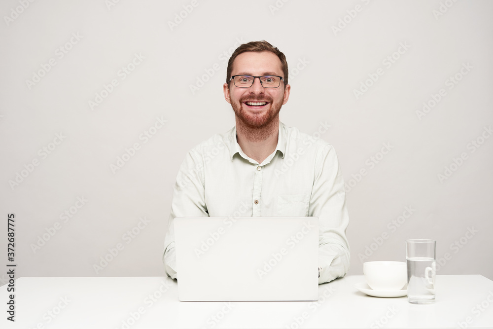 Happy young handsome unshaved male in eyewear looking gladly at camera with wide smile while working with his laptop over white background, being in high spirit Happy young handsome unshaved male in eyewear looking gladly at camera with wide smile while working with his laptop over white background, being in high spirit
