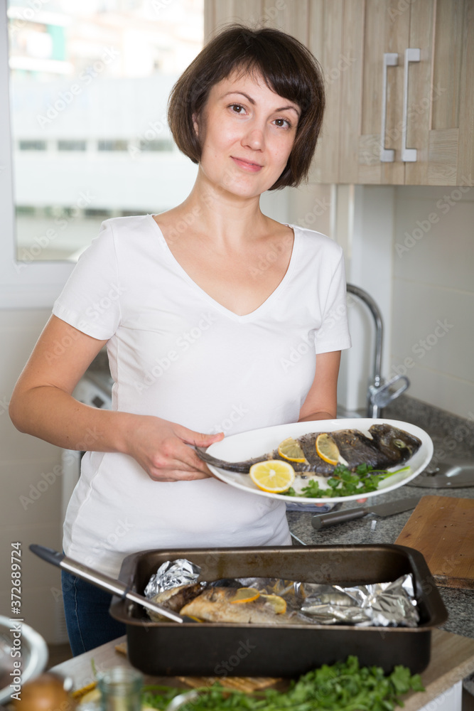 Positive housewife cooking dorado fish in baking dish at kitchen