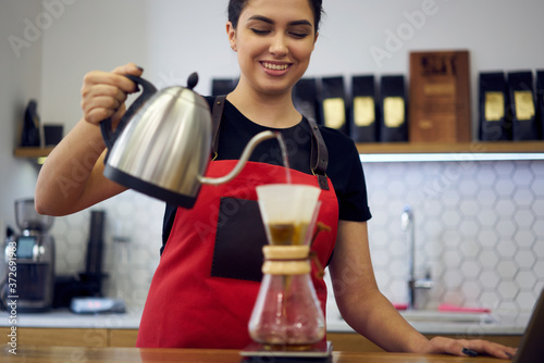 Cropped image of cheerful young woman working in own coffee shop and making tasty aroma coffee.Smiling barista holding kettle in hand and pouring water to preparing hot beverage for clients