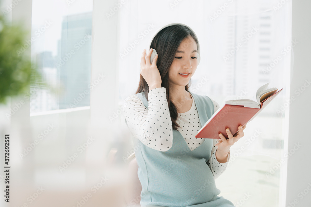 Pregnant asian woman listen to music with wireless headphone and reading a book to tell story to unborn child.