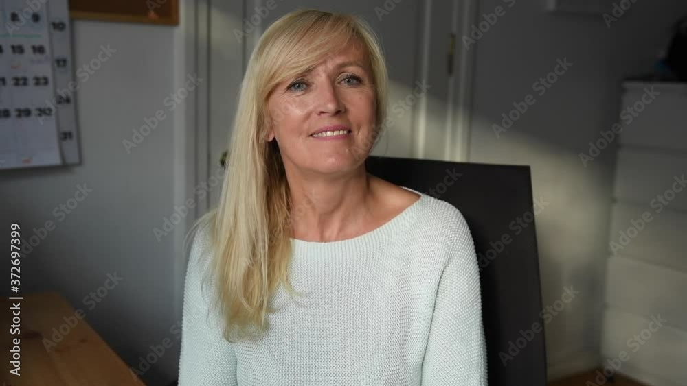 Portrait of smiling mature woman in her home office. Working from home businesswoman. Adult european female entrepreneur looking at camera sitting at her desk. Work video close up portrait indoors