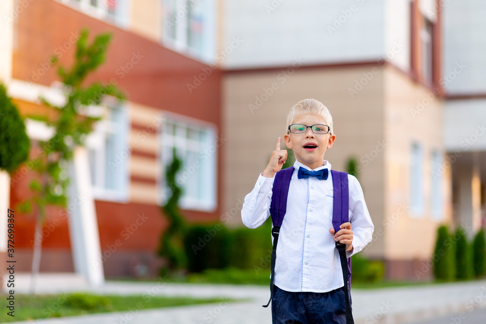 surprised boy schoolboy blond in glasses with a backpack stands at the school and shows a thumbs up. Day of knowledge surprised boy schoolboy blond in glasses with a backpack stands at the school and shows a thumbs up. Day of knowledge