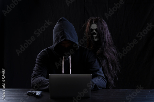 Canvas Print Mysterious man in the hood with skull inside using laptop on table with gun on isolated black background