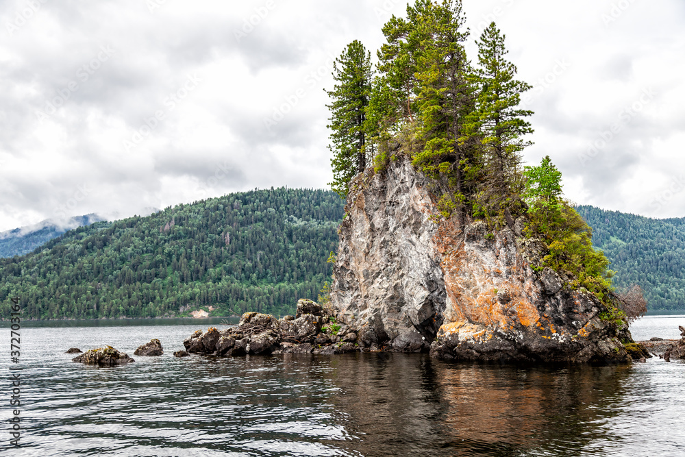 A rocky island with growing trees on it on the stones in the middle of the Teletskoye lake in Altai against the background of mountains in cloudy weather formed for centuries.