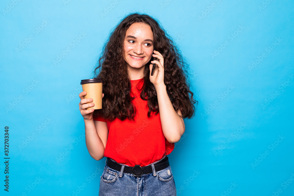 Young woman holding coffee to take away and talk mobile over isolated blue background