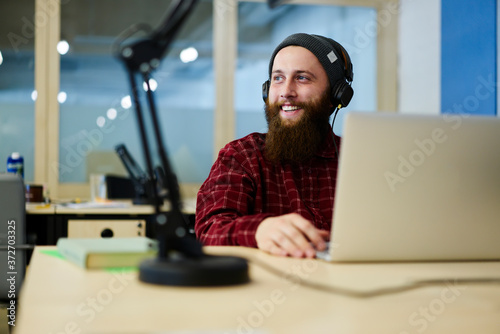 Cheerful graphic designer enjoying favorite compositions playing over cool headphones during work break.Bearded smiling copywriter listening good music while looking away sitting in office photo