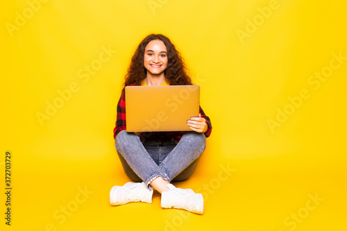 Portrait of a smiling young woman sitting cross-legged using a laptop on a yellow background