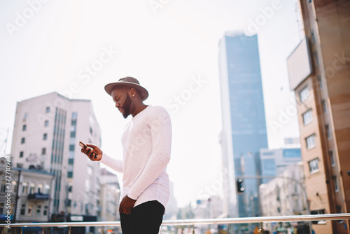 Young african american trendy dressed guy checking notification on cellphone standing on urban setting background, dark skinned male hipster guy using app on smartphone  and internet in roaming