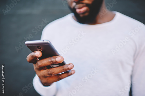 Cropped image of african american man holding modern smartphone dialing number, dark skinned male in white shirt using mobile phone for chatting in social networks via 4G internet connection outdoors